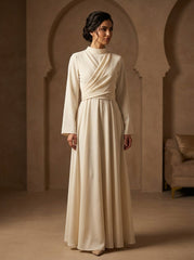 Woman wearing a long, light-colored dress in a warm-toned room with arched doorways.