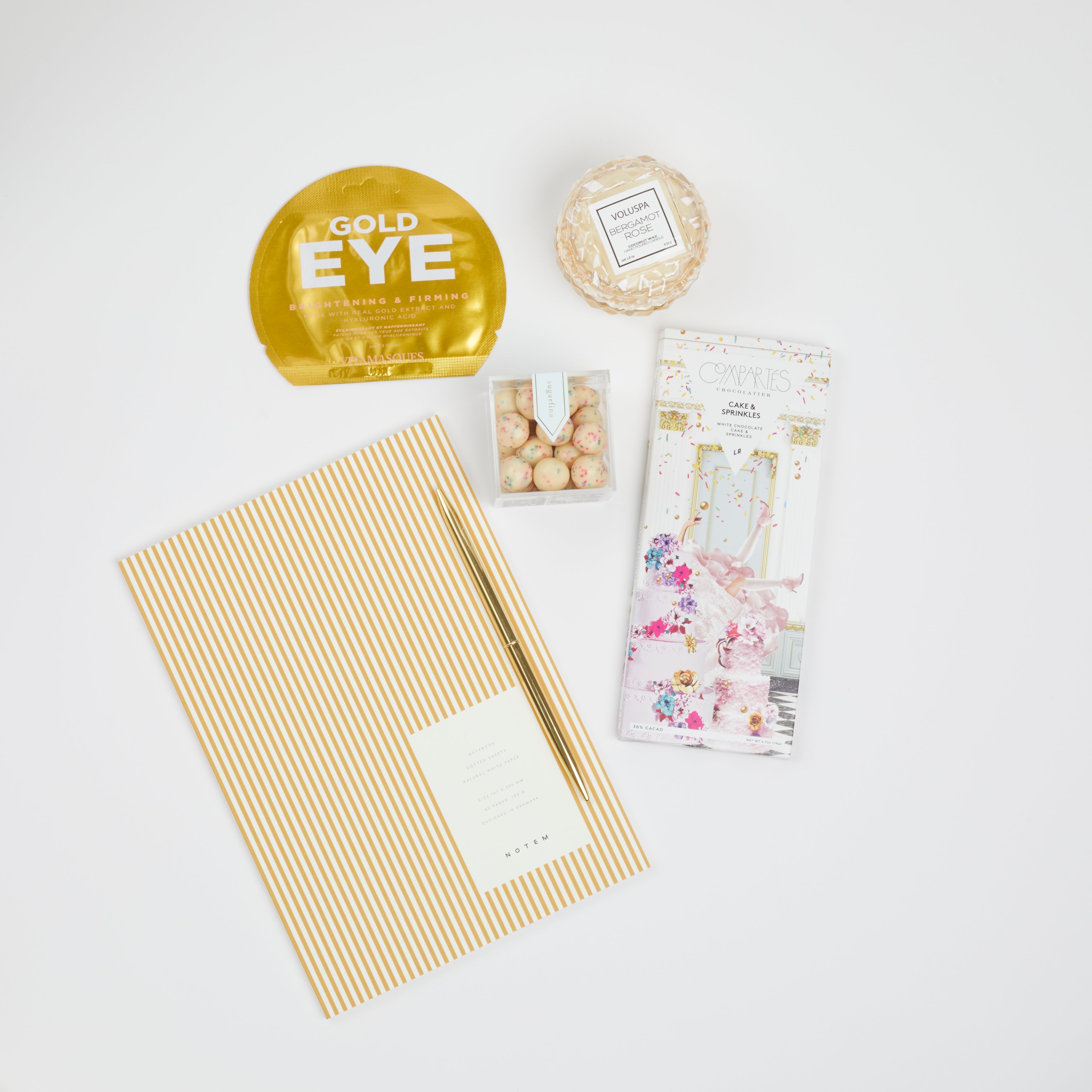 BOXFOX Gold Bullet Pen, Compartes Cake & Sprinkles White Chocolate Bar, Sugarfina Birthday Cake Cookie Bites, Voluspa Bergamot Macaron Candle and Vitamasque Gold Eye Mask. on white background