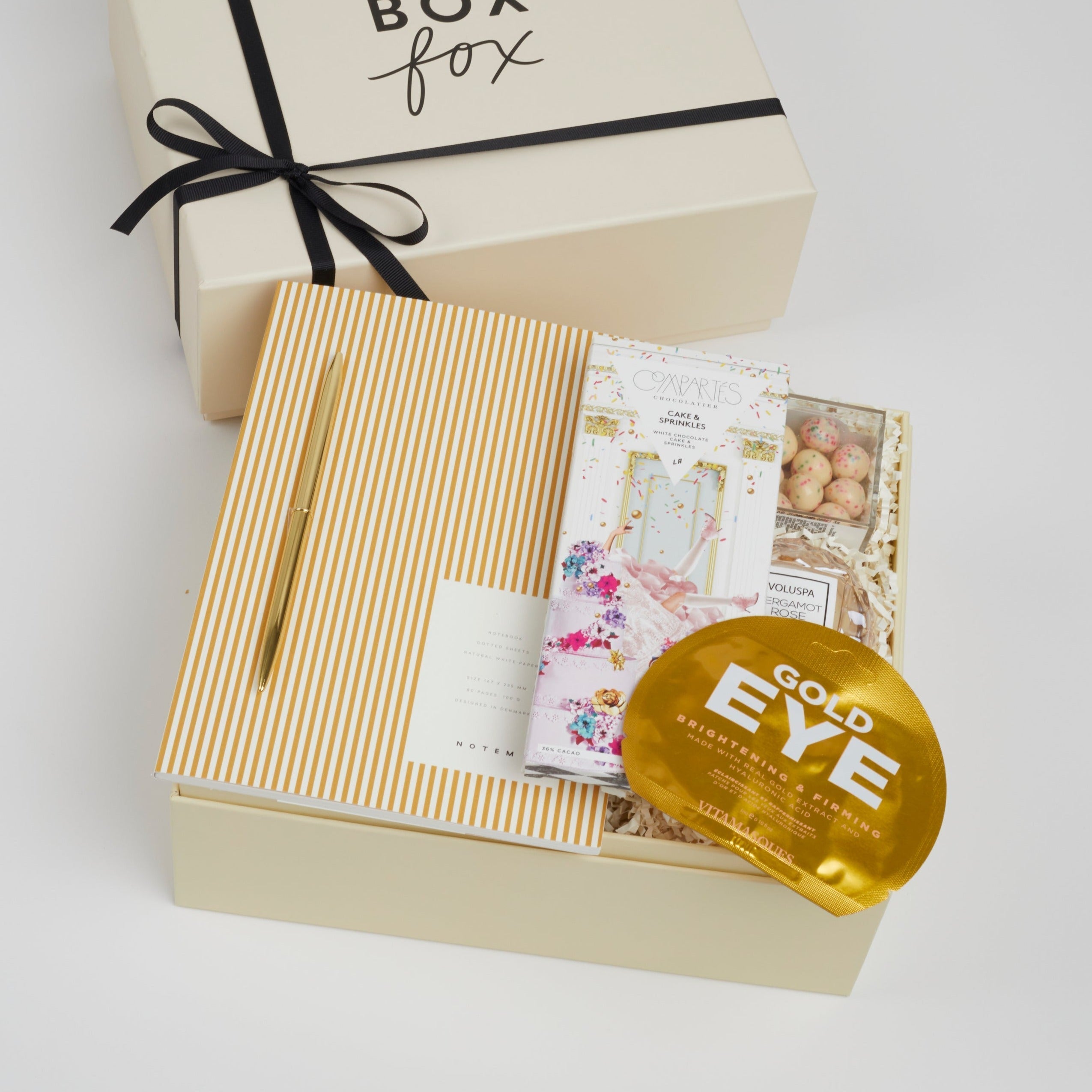 BOXFOX Creme Gift box containing Notem Gold Stripe notebook, BOXFOX Gold Bullet Pen, Compartes Cake & Sprinkles White Chocolate Bar, Sugarfina Birthday Cake Cookie Bites, Voluspa Bergamot Macaron Candle and Vitamasque Gold Eye Mask.